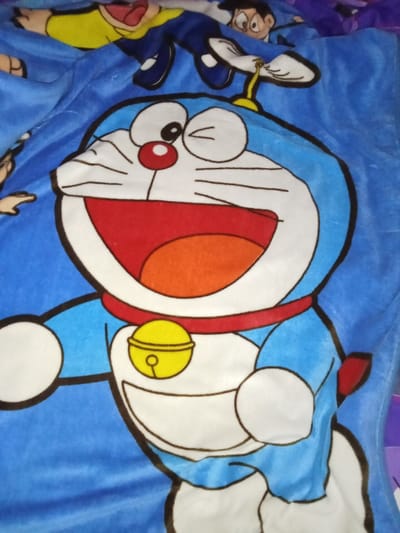 A plush blue fabric item, likely a blanket or duvet cover, is depicted, dominated by a large, colored illustration of the anime character Doraemon. Doraemon is centrally positioned, featuring a blue body, white face and belly, a red nose, black whiskers, and prominent white eyes. Its mouth is wide open, revealing a red interior and orange tongue/throat, conveying an expression of mirth. A red collar with a yellow bell is around its neck. Its left arm is extended to the viewer's left. In the upper background, partially visible illustrations of additional characters from the Doraemon franchise are present: one in yellow and blue attire on the upper left, and a head with dark hair on the upper right. The fabric exhibits a soft texture and minor folds. Lighting appears to be ambient indoor light. No visual evidence within the image defines a specific geographical location, including Inxuba Yethemba Local Municipality, South Africa.