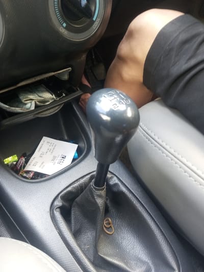 A car's interior is shown, focusing on the center console with a manual transmission gear stick. The gear stick has numbers indicating the gear positions. A rubber band rests near the base of the stick. Below the gear stick, a small storage compartment contains crumpled money, a receipt, and candy wrappers. A person's leg is visible near the seat.