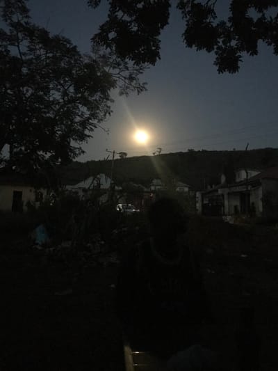 The image depicts a nighttime scene in Ushapa, Nigeria. A large, bright moon with a prominent lens flare illuminates the upper-center of a dark sky. In the lower-right foreground, a heavily silhouetted human figure, facing away from the viewer, is partially visible, showing an indistinct outline of the head and torso, appearing to wear a light-colored top. The immediate foreground consists of dark ground with some unidentifiable debris and a light blue object on the left. The midground features dense, dark vegetation and several dimly lit residential structures, including a white-walled building on the far left and additional structures across the center. A light-colored vehicle is faintly visible in the central-left amidst the foliage. Utility poles and overhead wires are discernible in the mid-right. The background consists of dark, undulating hills, silhouetted against the nocturnal sky. Dark tree branches frame the upper-left portion of the scene. No discernible actions or interactions are taking place.