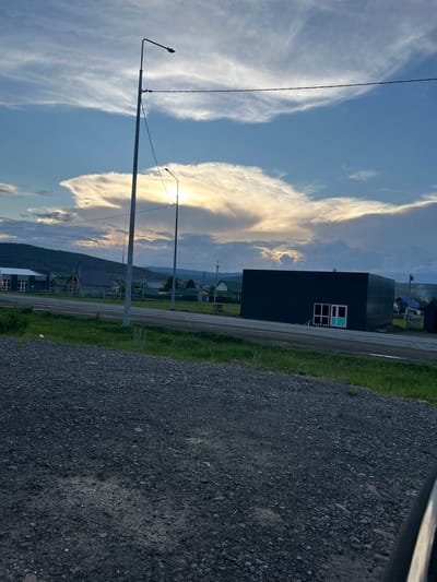 The image depicts a rural landscape in the village of Inzer, Russia, under a dramatic sky. In the foreground, a gravelly surface leads towards a grassy embankment. Beyond this, a paved road runs horizontally across the frame, separating the foreground from a collection of buildings and natural elements.

To the left, a modern, dark-colored building with white-framed windows is prominent. Further to the left, a lighter-colored building with a pitched roof is visible. The middle ground is occupied by a dark, rectangular structure with white-framed windows and a prominent turquoise door. To the right of this, a rural scene unfolds with smaller houses, some with green roofs, and fences.

The background features a rolling, tree-covered hill range under a vast sky filled with scattered, wispy clouds illuminated by a bright, diffused sunlight, suggesting either sunrise or sunset. Two tall lamp posts stand in the middle ground, one taller than the other, with power lines stretching between them. The overall impression is one of a quiet, somewhat remote settlement experiencing the atmospheric conditions of a dramatic sky.