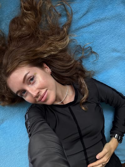 An overhead shot captures a person, likely female, lying on their back on a textured, light blue surface, resembling a blanket or towel. Their head is positioned towards the bottom-left of the frame, and they are looking directly at the camera with a faint, closed-mouth smile, suggesting a relaxed or self-assured expression.
The person has long, wavy, light brown hair that spreads out around their head on the blue background. They possess light blue eyes and defined eyebrows. A small facial piercing is visible above their upper lip, and a subtle yellowish-green mark is present near their left eye.
They are dressed in a long-sleeved black top, possibly athletic wear, which features dark, velvety or textured stripes across the torso and sleeves. Accessories include a thin silver-colored chain necklace, a black digital-style watch with silver accents on their left wrist, and a ring on a finger of their right hand, which rests on their chest. The scene appears to be an indoor setting in Ledukhi, Russia, though no specific architectural details or time of day are discernible. There is no visible text in the image.