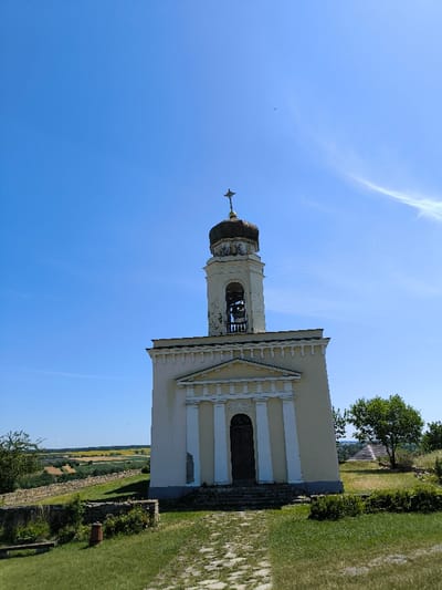 The image displays the St. Alexander Nevsky Church, situated within the Khotyn Fortress in Khotyn, Ukraine. The church is a light cream-colored, multi-story structure with a classical-style facade featuring white columns and a central dark wooden entrance door, accessed by a few stone steps. Above the main body, a multi-tiered bell tower rises, containing a visible bell within its open arched sections. The tower is topped by a dark, weathered dome, crowned with a golden Orthodox cross. Discoloration and wear are visible on the bell tower's upper portions.

In the foreground, a stone-paved path extends across a manicured green lawn towards the church entrance. To the left of the path, a low stone wall and green vegetation are present, with a small, reddish cylindrical object resting on the ground near the wall. The background on the left reveals a vast landscape of rolling green hills and distant cultivated fields, some exhibiting a yellow hue, under a clear, bright blue sky with minimal, faint cirrus cloud formations. To the right of the church, a single green deciduous tree is visible. No individuals or active events are depicted in the scene.
