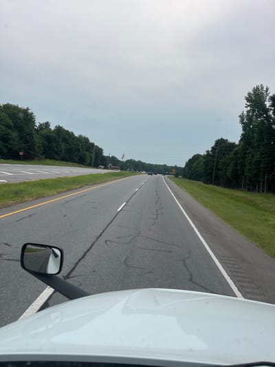 The image shows a perspective from the driver's seat of a white truck, looking forward down a two-lane highway. The road is paved and has white lane markings. There are trees lining both sides of the road. On the left side, there is a rest stop area with a sign that says "DO NOT ENTER". In the distance, there are a few cars on the road and an American flag flying. The sky is overcast. The location is Arcade, United States of America.