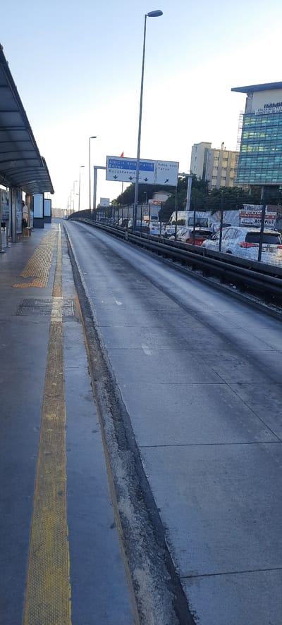 This image shows a street scene in Bahçelievler, Turkey. A wide, paved road stretches into the distance, bordered by a raised sidewalk with a yellow tactile paving strip. In the foreground, a section of a covered walkway or bus stop structure is visible, with some benches or seating areas inside. Across the road, beyond a metal railing, a parking lot filled with cars and a tall, modern building with a glass facade are visible.
Street signs are prominently displayed on a lamppost. One sign indicates directions to "Atatürk Hava" (likely Atatürk Airport), "Edirne," and "Küçükçekme." Another sign points towards "Bahçelievler Yanyol." The presence of these signs and the urban setting suggest this is a transportation hub or a main thoroughfare within the city.
The sky is clear and bright, suggesting it's daytime, likely morning or afternoon given the quality of light. There are no people prominently featured in the image, though there might be figures in the distance within the covered structure. The overall impression is one of a functional urban environment, possibly a transit area, with clear directional signage. The buildings and the road appear well-maintained.