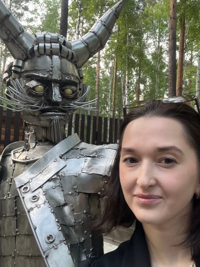 The image shows a woman taking a selfie in front of a large metal sculpture of a horned figure with a mustache, possibly a depiction of a character from folklore. The sculpture is made of many metal plates welded together, creating a segmented, armored appearance. The woman is in the foreground, smiling slightly and wearing a black jacket. Behind them, a wooden fence is visible, and beyond that, a forest with birch trees. The setting appears to be outdoors, in a natural environment. The location is specified as the Tal'tsy Museum of Wooden Architecture, Russia.