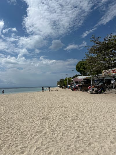 This image depicts a sandy beach scene with a calm ocean under a partly cloudy blue sky. In the foreground, the sand is textured with numerous footprints and small ridges. Several people are visible on the beach and near the water.
To the right of the frame, a row of small establishments with tin roofs and wooden structures lines the edge of the beach, suggesting a local market or vendor area. Signs on these structures indicate merchandise, with one visible sign featuring "TANDUAY" and "SADAG." Items like colorful umbrellas, life preservers, bags, and other goods are displayed outside these shops. Trees with green foliage are present behind these structures.
In the middle ground, the shallow, clear water gently meets the sandy shore. A few individuals are wading or standing in the water. Further back, the ocean extends towards a distant, hazy landmass. Along the far left of the beach, a few more people are seen walking or standing. The overall impression is of a casual, public beach area on a bright, possibly warm day. The scene is located in the city of Saavedra, Philippines.