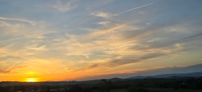 This is a photograph of a sunset over the village of Malo Buchino, Bulgaria. The sun is setting on the left side of the frame, casting a warm orange glow across the sky. There are several contrails from airplanes visible in the sky, stretching across the frame. The foreground shows a silhouetted landscape of rolling hills and trees. A few buildings are visible in the distance. The sky is a mix of blue, orange, and yellow hues, with wispy clouds scattered throughout. The overall impression is one of a peaceful and serene evening.