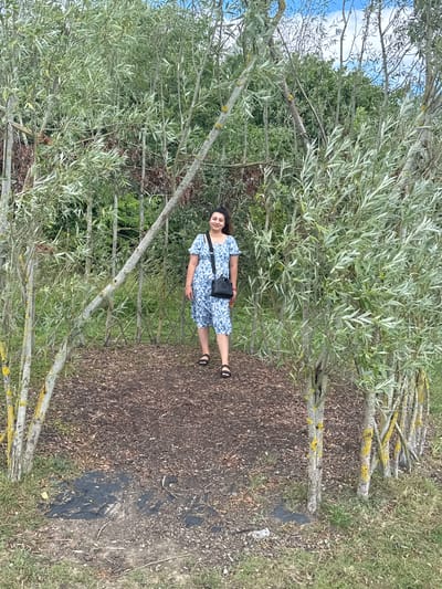 A woman stands in the middle of a clearing surrounded by a dense growth of trees and foliage. She is wearing a blue and white floral dress and a black crossbody bag. The ground in the clearing is covered with wood chips. The trees are slender and have long, narrow leaves. The sky above is partly cloudy. The scene appears to be a natural outdoor setting.