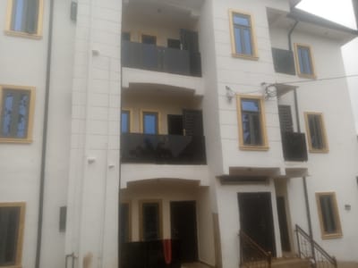 The image shows a multi-story white building with dark-framed windows, some with yellow trim. The building appears to be a residential or commercial property. Several balconies with dark glass railings are visible. There are also dark doors and some exterior lighting fixtures. The scene depicts a well-maintained building, suggesting it is located in a developed area. The overall impression is that of a modern structure within an urban environment.