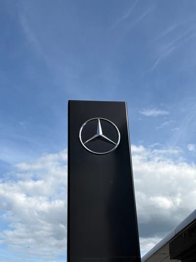 The image shows a Mercedes-Benz logo mounted on a black sign against a blue sky with scattered clouds. The sign appears to be attached to a building. It is not possible to confirm the location in the city of Riedlingen, Germany, from the image alone.