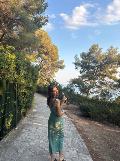 A woman in a green patterned dress stands on a stone path, holding a bottle, possibly beer. Lush greenery flanks the path, with trees overhead and a fence visible in the foreground. The sky is blue with scattered clouds. The photo is taken during the day, with warm lighting. The ground is made up of large grey flat stones. The woman is smiling.