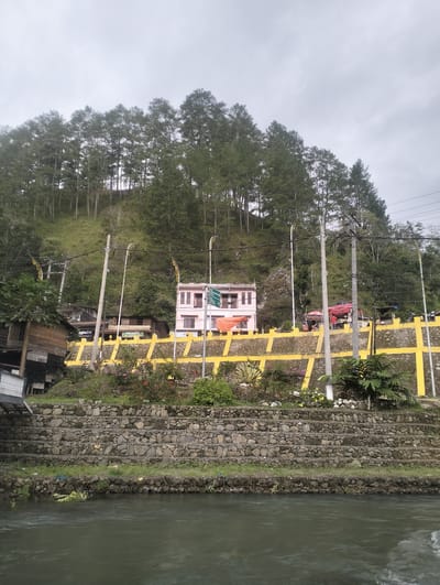 This media file depicts a roadside scene in a hilly, natural setting, likely in Blang Bebangka, Indonesia. The foreground is dominated by a river with murky, flowing water. Behind the river, a tiered retaining wall constructed from stones and wire mesh rises up the embankment, with sparse grass growing in the crevices.
Further up the slope, a yellow and grey patterned wall acts as a barrier along the road. Several buildings are visible, including a wooden structure on the left and a pastel pink building with multiple stories and balconies on the right, featuring arched windows. The pink building appears to have some commercial activity, with awnings and what looks like shops on the ground floor. A red car is parked on the upper level, with people gathered around it, suggesting a marketplace or roadside stop. Red and orange umbrellas are also visible, possibly for vendors.
The backdrop is a steep hillside covered in a dense forest of tall pine trees. The sky is overcast, indicating a cloudy or possibly misty day. Several utility poles with electrical wires crisscross the scene, suggesting the presence of modern infrastructure in this otherwise natural environment.
Notable visible text includes "ANGKAS 2" and "NASIGURIN" on signs of the buildings. The overall mood is serene and somewhat rustic, with a blend of natural beauty and everyday human activity. The time of day appears to be daytime, given the lighting conditions.
