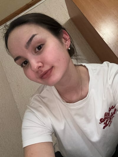 A young adult female is depicted in a close-up, chest-up shot. She has dark hair, dark eyebrows, and a neutral facial expression with a subtle smile, looking directly at the viewer. She is wearing a white short-sleeved t-shirt with dark red embroidered lettering, partially obscured, on the left chest area. A thin gold-colored necklace with a small pendant is visible around her neck. Her right ear has a small hoop earring with a clear stone in the lobe and a small cuff on the upper helix. The background behind her left side is a light-colored, textured surface, possibly a wall with a subtle pattern. To her right, a warm-toned, smooth wooden surface is visible, extending diagonally downwards from the upper right corner. The scene is located in Parizhskaya Kommuna, Russia.