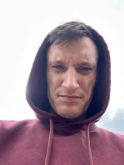 The image is a close-up, low-angle selfie of a man wearing a maroon hooded sweatshirt. The man has short, dark hair with some strands sticking up. His face is tense, with furrowed brows and a slightly downturned mouth. He has stubble on his chin and upper lip. The background is mostly a bright, overcast sky with some faint clouds visible on the right side. The lighting suggests it is daytime. The man's face and upper chest are the primary focus of the image. The location is stated to be Lysohirka, Ukraine, but this information is not visually verifiable from the image itself.