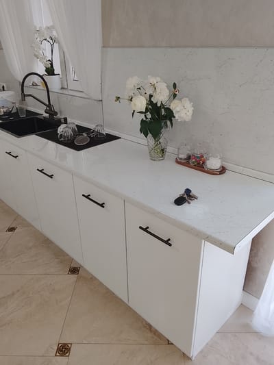 The image shows a kitchen interior. The kitchen has white cabinets with long, black, horizontal handles. A black double-basin sink is present, with a dark metal faucet. Dishes are drying on a black drying mat next to the sink. A vase of white flowers is on the countertop. To the right of the vase is a small wooden tray with three glass jars containing candy and other contents. A set of keys is lying on the countertop next to the tray. The flooring is a light brown tile with decorative corner accents. A window with white curtains is visible behind the sink. A white flower is on the windowsill. The walls appear to be a light marble pattern. The location is Assinovskaya, Russia.
