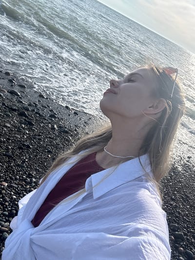 A woman stands on a black pebble beach at the edge of the water, likely in Gulripshi, Abkhazia, given the description. The woman is seen in profile, facing left. She wears a burgundy top beneath an unbuttoned, oversized white shirt. Sunglasses are perched on her head, and a thin, white beaded necklace adorns her neck. The beach consists of small, black pebbles, wet from the incoming waves. The sea is visible, shimmering with sunlight.