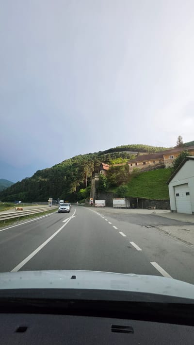 The media file captures a scene from a vehicle traveling along a winding road in Boița, Romania. The perspective is from inside the vehicle, looking out. The road curves to the right, with a lush green hillside to the left, covered in trees and some open grassy areas.

On the hillside, there are several structures. A large, light-colored building with a flat roof and garage-like doors is visible at the top of the incline. Further down, an older, ochre-colored building with multiple windows is situated on the slope. A smaller, rustic building with a red roof is nestled amongst the trees.

The road itself is asphalt with white lane markings. A silver car is driving ahead on the same road. The weather appears to be overcast, with a bright, hazy sky.

There are several signs visible along the side of the road. One sign has red and yellow markings and appears to be a traffic sign. Another rectangular sign has text, but it is not legible. Two white trucks are parked on the shoulder of the road near the signs.

Further down the road, a metal guardrail runs along the edge. A small herd of cows is visible grazing near the guardrail. The overall impression is of a scenic drive through a mountainous or hilly region. The time of day seems to be late morning or early afternoon, judging by the light.