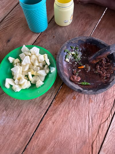 A top-down shot shows a wooden table with food preparation items in Blanggunci, Indonesia. On the left, a bright green bowl contains chopped white fruit, possibly pineapple. To the right, a dark, rough mortar and pestle holds a mixture of ground chilies, spices, and other ingredients, with two whole chilies, one green and one orange, visible. Behind the food items, a turquoise textured cup and a yellow plastic container with a cartoon illustration are present. A person's foot and leg are partially visible in the upper right corner of the image, suggesting they are seated at the table. The wooden table surface is aged with prominent grain patterns.