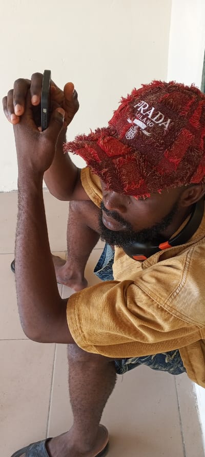 An individual is depicted crouching or squatting, observed from an elevated perspective in Ijotan, Nigeria. The subject is a male with dark skin, a black beard, and mustache. He wears a red, textured baseball cap with "PRADA MILANO" embroidered in white on the front, a mustard yellow short-sleeved collared shirt, and dark denim shorts. Black wireless headphones with orange accents are visible around his neck. He holds a black mobile phone horizontally with both hands, positioned in front of his chest and facing downwards, indicating engagement with the device. His arms are bare, and his legs are partially visible, one foot wearing a black slide sandal and the other appearing barefoot. The background consists of a plain light beige wall, and the floor is light brown tiled with visible grout lines.