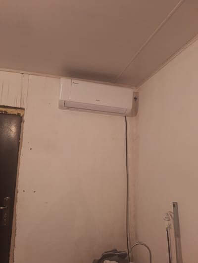 The image depicts an interior scene, likely in a residential setting, with an emphasis on an air conditioning unit mounted high on a white wall. The unit is a split system, with the indoor evaporator visible. It is white with a thin horizontal silver strip towards the bottom. The brand name "AUX" is clearly visible on the unit, and a smaller logo that appears to say "Inverter" is also present. A black power cord hangs vertically from the unit, running down the wall.
The wall itself is a light, textured color and appears somewhat aged, with visible staining and marks in places, particularly in the corner to the right of the air conditioner. Above the wall is a white ceiling with visible trim along the edge where it meets the wall.
To the left of the air conditioner, a dark-colored door frame is partially visible. The door itself is dark and has a silver handle. The area around the door frame appears to have been recently worked on, with visible foam insulation or sealant.
In the lower right portion of the image, a metal object, possibly a part of a cleaning tool or a fixture, stands upright against the wall. Below it, a portion of a sink faucet is visible, suggesting a bathroom or kitchen area. A pile of fabric, possibly clothing, is also present on the floor in the lower left. The overall environment appears somewhat utilitarian and possibly under renovation or in a state of disrepair.