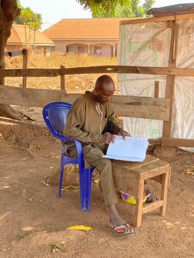 An adult male, with dark skin and a bald head, is depicted seated in Nev, Nigeria. He wears an olive green long-sleeved tunic, matching trousers, and black, white, and red striped flip-flops. The man is positioned on a blue plastic chair, leaning forward, holding a red pen in his right hand and an open light blue exercise book in his left hand. His left forearm rests on a small, rustic wooden stool that serves as a writing surface. Underneath the stool, a colorful plastic bowl (yellow, purple, green) is partially visible. The immediate ground consists of dry, reddish-brown earth with scattered dry leaves and exposed tree roots. To the man's right, a makeshift structure is present, constructed from wooden planks and partially covered with white, repurposed sack material bearing faint red and green branding. In the background, under bright daylight, an expanse of dry, light-colored vegetation leads to two single-story buildings featuring light orange corrugated metal roofs. One building exhibits visible windows and suspended laundry.