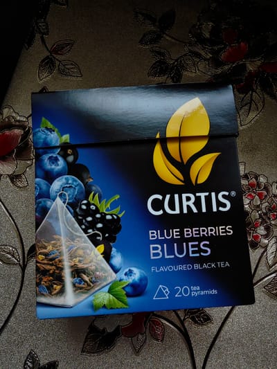 The image shows a close-up of a box of Curtis Blue Berries Blues flavored black tea. The box is black with a blue gradient and features images of blueberries, blackcurrants, and a single tea pyramid bag. The brand name "CURTIS" is prominently displayed in white lettering. Below the brand name, the text "BLUE BERRIES BLUES" and "FLAVOURED BLACK TEA" are visible. At the bottom of the box, the text "20 tea pyramids" is shown next to a graphic of a tea pyramid. The box is placed on a patterned surface, possibly a tablecloth or a textured background. The overall lighting suggests an indoor setting. There is no indication of the city of Dubovoye, Russia in the image.
