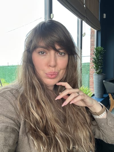 A woman with long, wavy brown hair is posing for a selfie. She is wearing a brown shirt and has a ring on her left hand. Behind her is a window with a view of a green field and a brick building. There is a plant in a gray pot on the windowsill. The location appears to be indoors, possibly a cafe or home in Marneuli, Georgia, based on the type of buildings and vegetation visible through the window.