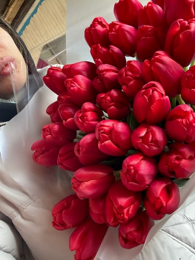 A close-up image depicts a large bouquet consisting of approximately 40-50 vibrant red tulips, mostly closed or partially open, wrapped in white material. A person's lower face, including the mouth and chin, is visible on the upper left, partially obscured by a clear plastic face shield. Dark hair is visible near the top of the person's head. A light-colored puffer jacket is visible on the left and bottom, worn by the individual. The background on the upper left consists of a light brown wooden structure with horizontal lines, indicating an indoor setting in Trudobelikovskiy, Russia.