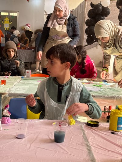 The image captures an indoor art and craft session occurring in Kafr Batna, Syria. In the foreground, a young boy with dark hair and fair skin is seated at a table covered with a pink plastic tablecloth. He wears a teal long-sleeved shirt and a translucent plastic apron. His head is turned to the left, and he holds a spoon containing a yellow substance in his right hand, while his left hand manipulates a yellow-colored, irregularly shaped object on the table. To his left on the table are two clear plastic cups; one is mostly empty with purple residue, and the other contains a blue liquid with red specks on its rim. A yellow paint jar with its black lid is also present on the table to his right. A white figurine with purple details stands on the table to his left.
Behind and to the left of the foreground boy, another boy wearing a brown hooded jacket is seated, looking down at a mobile phone in his hands. A clear plastic cup with red liquid and a small red and white box are on the table in front of him.
An adult woman, wearing a dark jacket, a light pink hijab, and a beige apron with a graphic design, stands to the left of the foreground boy, observing the activity. To the right, another adult woman, dressed in a light beige hijab and apron, leans forward over an adjacent table, engaged in a task; a bracelet displaying green, white, and red stripes is visible on her right wrist. A child in a pink jacket is partially visible behind the foreground boy, also participating in an activity.
The background reveals several other individuals, including adults and children, seated at tables covered with light-colored plastic sheets, all engaged in creative tasks. The room features light-colored walls. Decorations include a cluster of black and white balloons on the right and a yellow robot-shaped cutout with the letters "SET" displayed on the far-left wall.