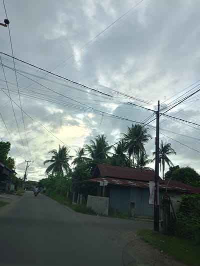 The image depicts a street scene in Meunasahteungkudigadong, Indonesia, under an overcast sky. A paved road curves slightly, with a lone motorcyclist visible in the distance. On the right side of the road stands a dilapidated building with a corrugated metal exterior and a rusty red roof. Lush tropical vegetation, including palm trees, surrounds the building and lines the roadside. A utility pole stands near the building, draped with power lines that crisscross the sky. A sidewalk runs along the edge of the road.