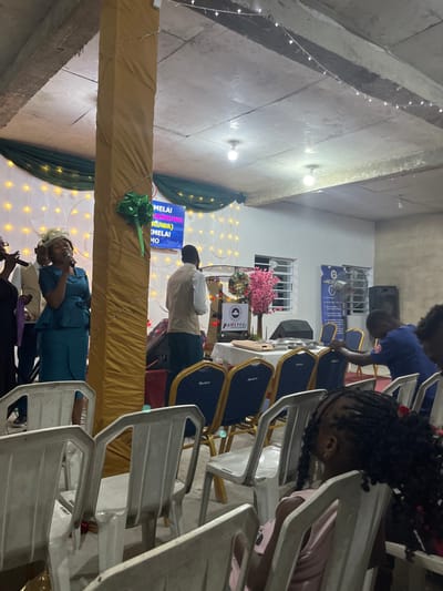 The interior shows people seated on plastic and upholstered chairs in a brightly lit space. A stage is adorned with gold fabric, lights, and greenery. A screen displays text. A podium with flowers and a logo is visible, suggesting a religious gathering or performance. A woman in a blue outfit is speaking into a microphone. There are a few children at the foreground.