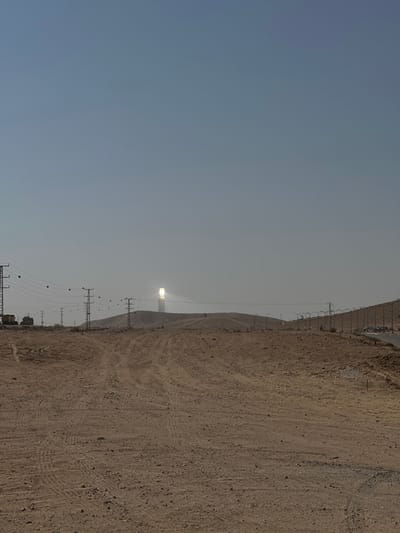 A landscape shows a stark contrast between the clear, light blue sky and the arid, brown ground. A solar tower with a bright glare stands out against the horizon. Power lines stretch along the side, indicating human infrastructure within this dry environment. The ground is marked with tire tracks, possibly from vehicles driving across the terrain.