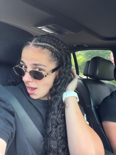 A young woman with braided hair and sunglasses is sitting in the passenger seat of a car. She is looking at the camera with her mouth slightly open. The car appears to be in motion, as indicated by the blurry trees visible through the window. The scene is set in Брусовeте, Bulgaria.