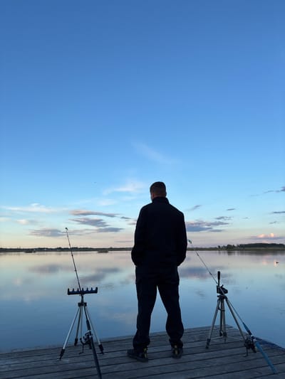 A low-angle, horizontally-oriented photograph captures a man fishing from a wooden dock in Khryashchevka, Russia. The sky is mostly clear with some wispy clouds. The man, seen from behind, is wearing dark clothing and stands between two fishing rod setups. Each setup includes a tripod holding a fishing rod over the water. The water is calm, reflecting the sky and distant trees. The wooden dock is composed of planks, and the overall scene appears to be at dusk or dawn, given the soft lighting.