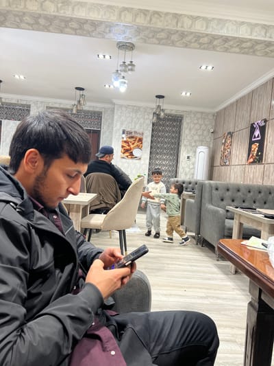 An indoor shot shows a man focused on his phone, seated near two children running past. The scene unfolds in a restaurant featuring gray tufted furniture and light walls. A sign with french fries is on the wall, and posters hang above. Additional people are seated in the background, contributing to the lively yet relaxed atmosphere.