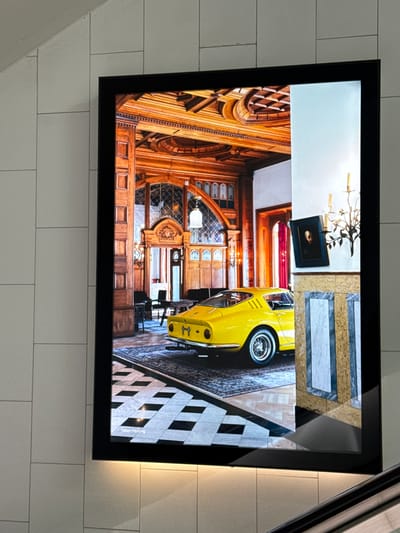 The image shows a large digital display mounted on a tiled wall. The display features a photograph of a vintage yellow sports car parked inside a grand, wood-paneled room with a high ceiling. The room has arched windows, intricate woodwork, and a patterned rug on the floor. A black and white checkered floor pattern is visible in the foreground of the photograph. The display is illuminated, and there is a subtle glow from a light source below it, possibly from an escalator handrail. The background wall is made of light-colored square tiles. The photograph within the display has a small text overlay in the bottom left corner that reads "Valet Parking". The location is specified as Sesto San Giovanni, Italy.