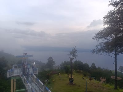 A wide shot captures a scenic overlook on a hazy day, overlooking a vast body of water. In the foreground, a modern observation deck, constructed with a white railing and blue accents, extends over a lush green hillside. Several individuals are visible on the deck, some standing and observing the view, others posing for photos. A sign with blue text that reads "SINARJARUNJUNG SARABEL" and a heart symbol is mounted above the deck. The hillside is dotted with trees and scattered with various decorative elements, including a crescent moon sculpture, a set of wooden benches, and what appears to be a playground structure with an animal figurine. A large, mature pine tree stands prominently on the right side of the frame, its branches reaching towards the overcast sky. In the background, the expansive water stretches towards a distant, hazy horizon. The location is identified as Nagaritongah, Indonesia.
