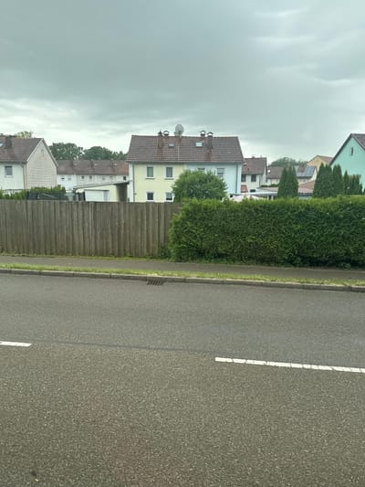 The image depicts a street scene in Memmingen, Germany. In the foreground, a wet asphalt road with a white dashed line is visible, indicating a lane divider. A concrete curb separates the road from a grassy verge. Behind the verge, a tall, weathered wooden fence runs horizontally across the frame, partially obscured by a dense, well-maintained green hedge.
Beyond the hedge and fence, several residential buildings are visible. These include houses with pitched, tiled roofs and light-colored facades. One house in the center is painted a pale yellow and has multiple windows. Another house to the left has a white facade with a patterned texture. Further back, other homes with varying rooflines and colors can be seen, along with some trees. The sky is overcast and appears a uniform gray. There are no people or animals visible in the image. The overall impression is a quiet residential area, possibly on a cloudy or rainy day.