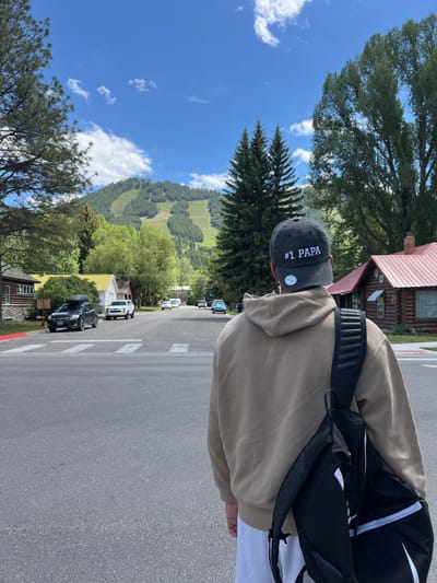 A person wearing a tan hoodie and a black baseball cap with the words "#1 PAPA" embroidered on it is standing on a street in Jackson, Wyoming, United States of America. The person is seen from behind, with a black backpack slung over their shoulder. In the background, there is a street lined with houses and trees, with a ski slope visible on a mountain in the distance. Several cars are parked on the street and in driveways. The sky is blue with some white clouds.