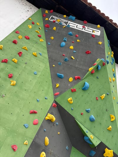This image depicts a section of an outdoor climbing wall, likely located in Баня, Bulgaria. The wall is constructed from multiple angled panels, predominantly colored matte green and dark grey. These panels are affixed with numerous artificial climbing holds in various shapes, sizes, and colors, including bright red, vivid yellow, and light blue. The holds are arranged across the surfaces, creating routes for climbing. A white logo, "EL CLIMB," with a distinctive 'X' symbol, is prominently visible on one of the dark grey upper panels. The wall is positioned against a textured white building exterior on the left. Above the climbing structure, a dark wooden beam and tile roof structure is visible, suggesting the climbing wall is integrated into a building or covered by an overhang. There are no people present, nor any discernible actions or interactions occurring in the scene.