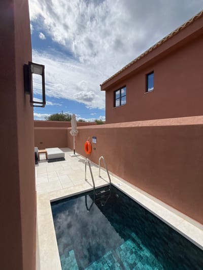 The image displays an outdoor private swimming pool and patio area, located in the Archanes Municipal Unit, Greece, under a partly cloudy blue sky.

The foreground on the left features a terracotta-colored stucco wall with a black, open-frame outdoor light fixture mounted vertically. A rectangular swimming pool, lined with dark teal-green mosaic tiles, occupies the lower center and right, reflecting the sky. Stainless steel handrails are installed at the visible end of the pool, which has a white coping. A "1.30m" depth marker is present on the coping near the pool's edge.

Behind the pool, a light-colored tiled patio contains a white, modern sun lounger with a wooden base and a folded white patio umbrella on a white stand. This patio is enclosed by a continuous terracotta-colored stucco wall. Mounted on this wall, above the pool, is a bright orange lifebuoy and a small, illegible informational sign.

To the right, a two-story building with matching terracotta-colored stucco walls and a traditional clay tile roof is visible. It features two dark-framed windows, one with multiple panes, reflecting the sky. In the distant background, above the perimeter walls, sparse green foliage and utility lines are visible. No individuals are present in the scene.