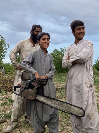 The image captures three males—an adult man and two younger individuals—standing outdoors in a rural setting, likely involved in land clearing or logging activities in Qila Saifullah, Pakistan.

The man, positioned to the left and slightly behind, has a beard and wears a light-colored traditional shalwar kameez and a patterned cap. He is smiling warmly. In the center, a young boy with a bright smile wears a grey shalwar kameez and a similar cap. He holds a large, heavy-duty chainsaw with both hands, its long blade extending downwards. To the right, another young man or older boy, also wearing a light-colored shalwar kameez and cap, stands with his arms crossed, looking towards the right with a contented expression.

All three subjects appear joyful and are dressed in traditional South Asian attire, with their clothes showing signs of dirt and wear, consistent with manual labor. The background consists of green trees and foliage under a cloudy, overcast sky, indicating daytime. The ground is uneven and earthy, with what appears to be cut branches or debris on the left side of the frame. There is no visible text on clothing or the chainsaw.