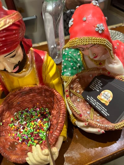 The image shows two figurines, a man and a woman, on a wooden table in a restaurant in Saint-Josse-ten-Noode, Belgium. The man is wearing a yellow and red turban and a yellow and red robe. He is holding a basket filled with colorful candies. The woman is wearing a red and green sari and holding an empty basket. A business card for a nearby Indian restaurant, "RANGLA PUNJAB", is sitting on the table. A clear plastic bottle with a white cap is also visible in the background. The table surface is brown and wooden. The overall ambiance is that of a restaurant, with the figurines serving as decorative elements.