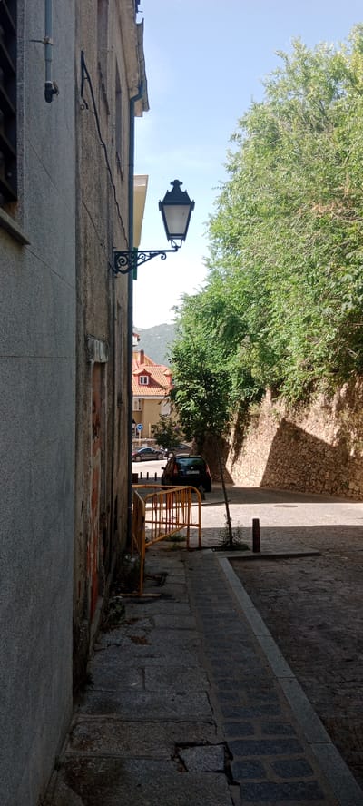 A sunlit, narrow street in San Lorenzo de El Escorial, Spain, is captured in a vertical photograph. The scene is dominated by a building's textured grey facade on the left, adorned with an ornate black street lamp attached to an iron bracket. The lamp, with its classic design, hangs over the cobbled sidewalk. 

Beyond the building, a glimpse of a building with a red-tiled roof and white window frames suggests a residential area. Further down the street, a dark-colored car is parked, and another dark car is visible in the distance. A yellow metal barrier, possibly for construction or traffic control, is positioned along the sidewalk. 

Lush green trees and foliage spill over from the right side of the frame, bordering a stone wall that rises behind the street. The dappled sunlight creates a pattern of light and shadow on the ground, indicating it is daytime. The sky is a clear, pale blue. The overall impression is one of a quiet, historic European street on a pleasant day. No people are visible in the image. The license plate of the parked car reads "5915 DTW".