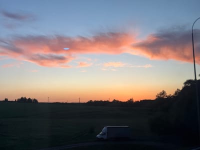 The image shows a twilight scene in Федькино, Russia. In the foreground, there is a parked white delivery truck on a dark road. A field stretches out behind it, leading to a line of trees and utility poles in the mid-ground. The sky is multi-colored, transitioning from blue at the top to orange near the horizon, with pink clouds visible. A streetlight pole is visible on the right edge of the frame.