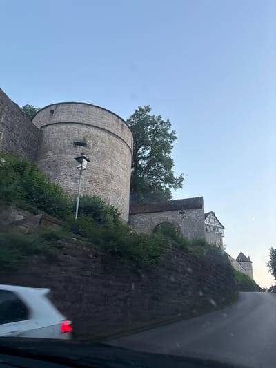 The image shows a road curving uphill towards the historic town of Vellberg, Germany. On the left, a large, circular stone tower with a small window is visible, part of the town's medieval fortifications. The tower is surrounded by lush green foliage and a stone wall. Further up the hill, an arched stone gateway leads into the town, with a half-timbered building perched above it. To the right, a conical-roofed tower can be seen in the distance, along with more of the town's architecture. The sky is a clear, pale blue with a faint crescent moon visible. In the foreground, the rear of a white car is visible, with its brake lights illuminated. The scene appears to be captured at dusk.