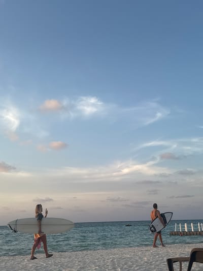 Two people are holding surfboards on a beach. One person is holding a smaller surfboard, while the other person is holding a larger surfboard. The beach is located in Himmafushi, Maldives. The sky is blue with some clouds.