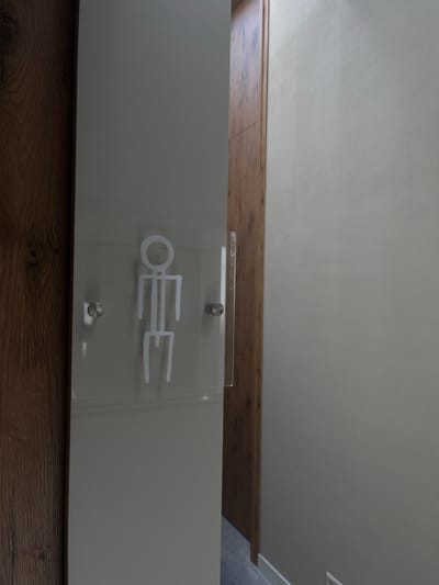 The image displays an interior scene in Overveen, Netherlands, centered on a light gray door with an attached restroom sign. The sign is a clear rectangular acrylic panel, approximately 30-40 cm high, held in place by two silver-toned stand-off fasteners, one on each side. A white, stylized stick-figure pictogram, consisting of a circular head, rectangular torso, two horizontal arm segments, and two downward-extending leg segments with outward-pointing feet, is etched or printed on the acrylic. The light gray door is slightly ajar, revealing a narrow brown wooden door frame or wall section directly behind its right edge. To the far right of this, a plain light beige wall is visible. To the immediate left of the light gray door, a wider section of brown wooden wall or door frame, exhibiting a distinct grain pattern, fills the left portion of the image. The area visible through the slightly open door appears darker, suggesting a recessed hallway or further interior space. No individuals, actions, or interactions are depicted.