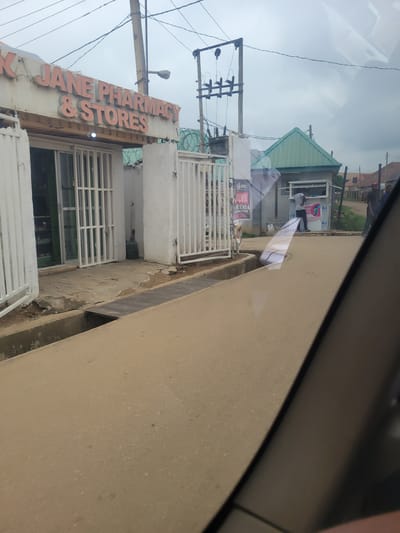 The image depicts a street scene in Katambe, Nigeria. In the foreground, a tan-colored road with scattered debris occupies the lower half of the frame. A concrete drainage channel runs along the edge of the road. To the left, a white building with metal security gates is visible. Above the entrance, a sign reads "AJANE PHARMACY & STORES" in orange lettering. Inside the pharmacy, shelves stocked with various items can be seen through the glass. Adjacent to the pharmacy, a similar white building with white metal gates extends further. Further down the street, to the right, a smaller green-roofed structure serves as a kiosk or small shop. A person in a white shirt is visible standing in front of this kiosk, seemingly interacting with it. Other people are present in the background, some walking and others standing near buildings. Electrical poles with wires are prominent in the sky. The overall environment appears to be a commercial area with a mix of residential and business establishments. The sky is overcast.