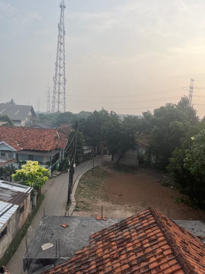 The image depicts a hazy, overcast day in Pasir Gadung, Indonesia. The scene is dominated by a tall telecommunications tower in the distance, partially obscured by the atmospheric conditions. In the foreground, a residential area with red-tiled roofs is visible, along with a narrow road and surrounding vegetation. Power lines crisscross the sky, and electrical pylons can be seen in the mid-ground. The overall impression is one of a somewhat undeveloped urban or semi-urban environment. No people or specific actions are clearly identifiable in the image.